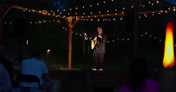 Male musician performing acoustic set under pergola with string lights at night