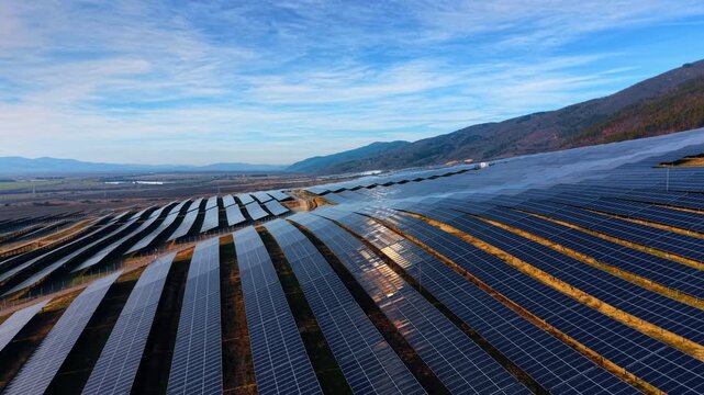 Solar panel rows on mountain hillside. Converging lines of photovoltaic arrays under wispy clouds with valley plains beyond.