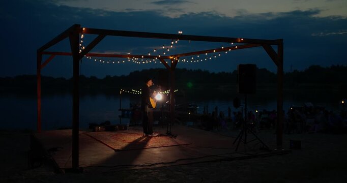Solo guitarist performing on illuminated wooden lakeshore stage at dusk