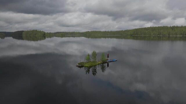 Drone footage of a pristine lake in Mujej&auml;rvi, North Karelia, Finland featuring a tiny island, a solitary kayaker, and vast coniferous forests under a moody sky.