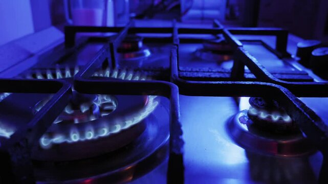 Male hand lights a gas stove burner with a flaming match, igniting blue flames. Close-up shot in a dark kitchen with dramatic blue ambient lighting and smooth camera movement.