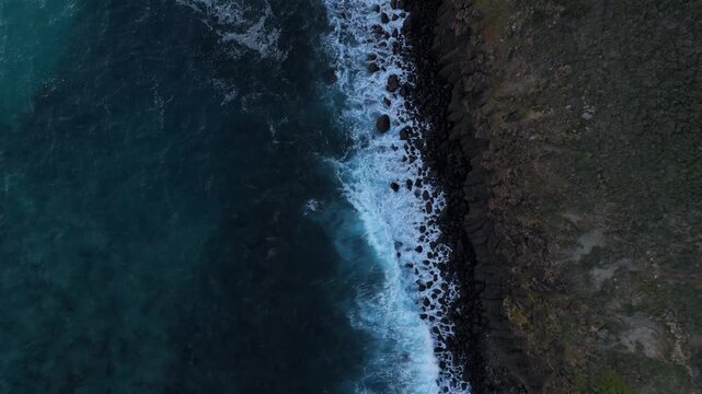 Aerial drone top-down view pulling up to reveal a wider landscape of ocean waves crashing against dark rocks at Pyramid Rock, Australia.