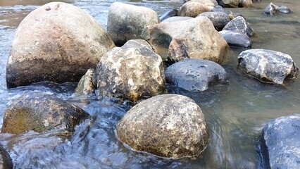 Fototapeta premium Large smooth river boulders surrounded by shallow flowing water. Natural rounded stones with textured surfaces create an organic background, showcasing erosion, geology, and the beauty of nature.
