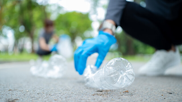 Close-up hand in a blue glove picking up a plastic bottle from a park path. Blurred volunteer in background. Concept of environmental conservation, plogging, and recycling.