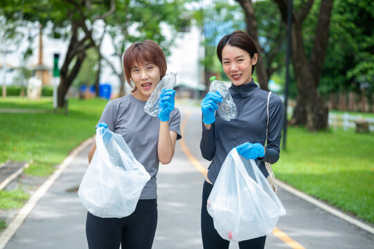 Two happy Asian volunteers in activewear holding bags and showing collected plastic waste in a scenic park, ideal for representing plogging and conservation.
