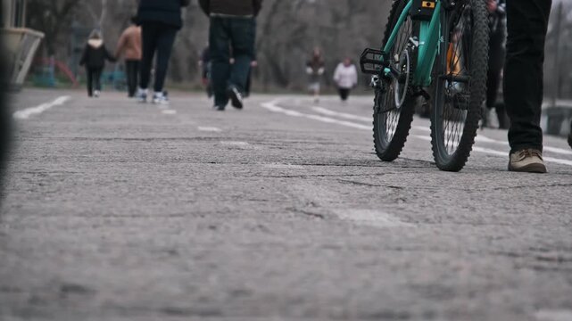 A low-angle shot captures a bicycle and people walking on a paved path in an urban park. The blurred foreground and ground-level view emphasize the movement of the wheels.