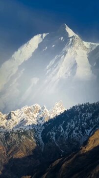The majestic, snow-covered peak of Rakaposhi rises dramatically above the rugged, forested slopes and deep, shadowed valleys of the Karakoram range in northern Pakistan, illumin snow covered mountains