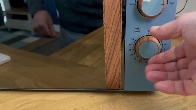 Closeup of a man&rsquo;s hands closing the door of a microwave oven, with a container of food inside to be reheated on the turntable, then setting the timer control to activate it.