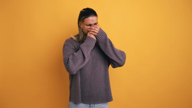 Young man with long hair feeling disgusted by an awful smell, covering his nose and mouth with his hand on a vibrant yellow background