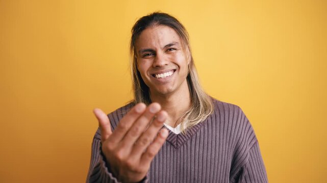 Handsome young man with long hair smiling and making an inviting come here gesture with his hands. Positive emotions in a studio setting