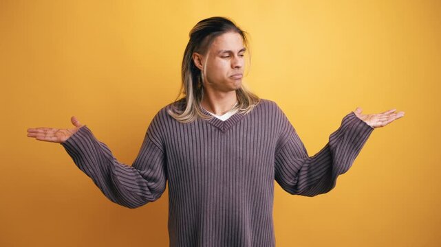 Unsure young man with long hair shrugging his shoulders and spreading his arms, expressing confusion and doubt against a yellow background