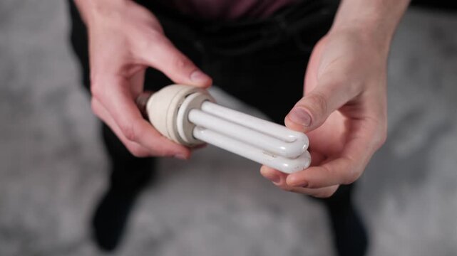 Close-up of male hands holding and inspecting an old compact fluorescent light bulb. The used bulb shows signs of wear to emphasize sustainability and recycling needs.