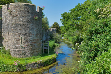 An ancient stone castle wall with a round turret stands beside a narrow moat. Lush green trees and bushes line the water's edge - The Bishop's Palace & Gardens - Wells - Somerset - UK © Robert