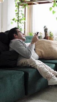 Young man with a physical difference sitting comfortably on the sofa, smiling while playing a videogame on his smartphone in his apartment