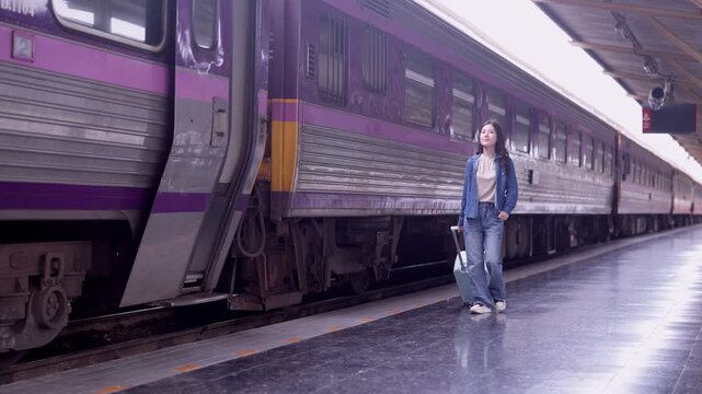 Young Asian woman traveler waiting on empty train platform with luggage beside purple passenger train in modern railway station