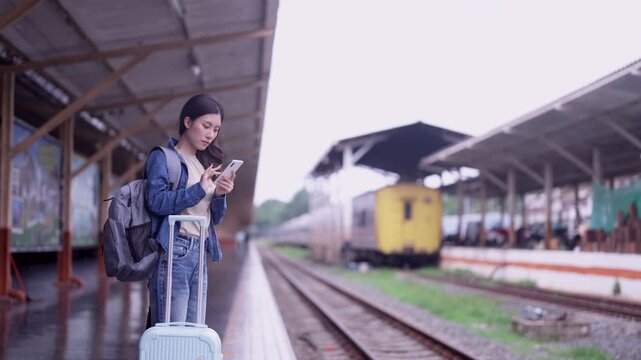 Young Asian woman traveler waiting on train platform with rolling suitcase and backpack checking smartphone under cloudy sky at vintage railway station