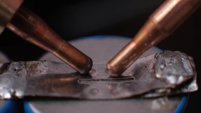 Extreme close-up slow motion of copper electrodes welding a nickel strip to a lithium battery. Subtle smoke rising from the contact point emphasizes the heat of the welding process.