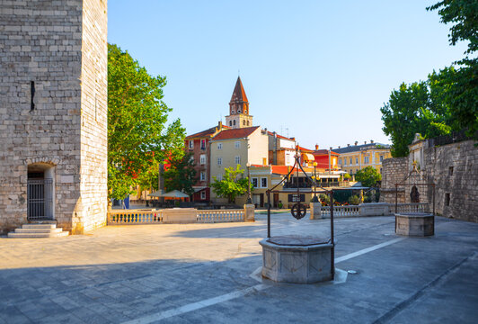 Five Wells Square in Zadar, Croatia, features historic stone wellheads aligned on a wide, sunlit public plaza. Medieval Captain Tower and distant church spire frame this idyllic Dalmatian setting