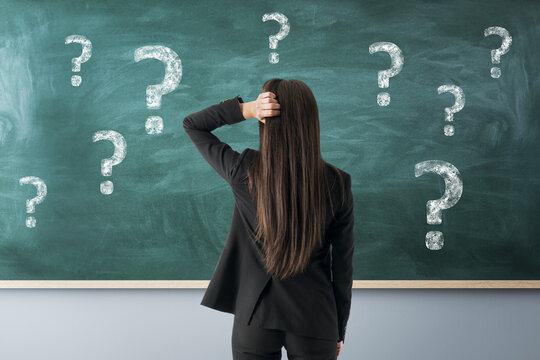 Confused woman standing before chalkboard filled with question marks, reflecting uncertainty and problem solving in educational setting.
