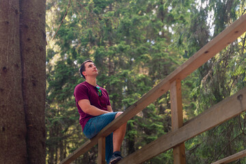 Fototapeta premium Young man resting on a wooden railing in green forest, wearing casual clothes and looking upward near a large tree with soft natural light and blurred