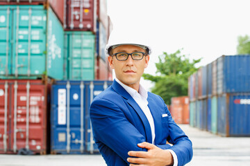 Fototapeta premium Serious logistics professional stands confidently before stacked shipping containers wearing blue suit and white hard hat, exhibiting focused demeanor during industrial operations..