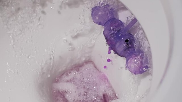 Super slow motion close-up of a toilet bowl flushing water over a purple rim block. The slow formation of violet bubbles highlights the effectiveness of the sanitation.