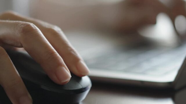 4K Video close up of a businesswoman hand scrolling and clicking a wireless computer mouse while working on a laptop at a desk, professional office employee typing in the blurred background.
