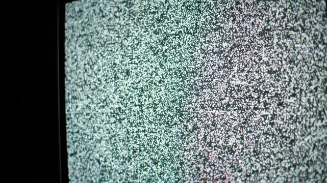 Close-up angled perspective displays a flickering white and black static noise pattern on an old CRT television screen. The no signal interference creates a distortion effect.