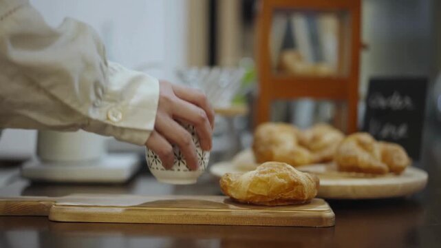 A barista serves a fresh croissant with blue tongs onto a wooden platter, adds a patterned ceramic cup, and pours black coffee from a glass server in a cozy cafe, shot in 4K Video.