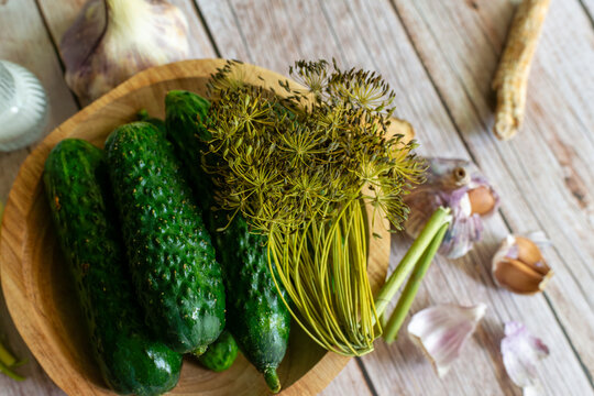 Fresh pickling cucumbers lie in a wooden bowl with dill seed heads, garlic cloves, horseradish root, and pale wooden boards for homemade preserving.