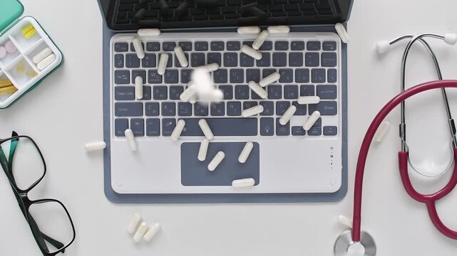 White capsules fall onto laptop keyboard beside stethoscope, showing online pharmacy, telemedicine and digital health work