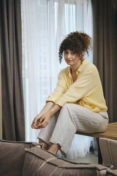 Cute woman with curly hair sits casually on a table in a well-lit room. She wears a yellow shirt and grey loose pants, giving off a relaxed vibe