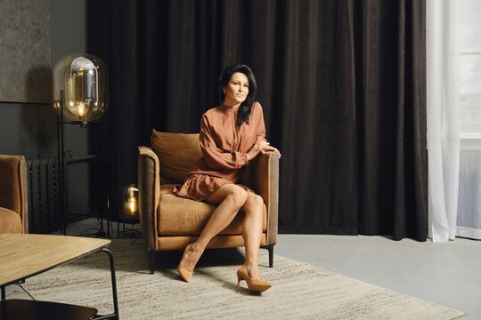 A woman with long black hair sits gracefully in a stylish brown armchair. She wears a fashionable rust-colored dress and light-colored heels