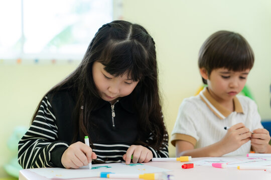 Focused little Asian girl drawing with markers next to her classmate during art lesson in kindergarten