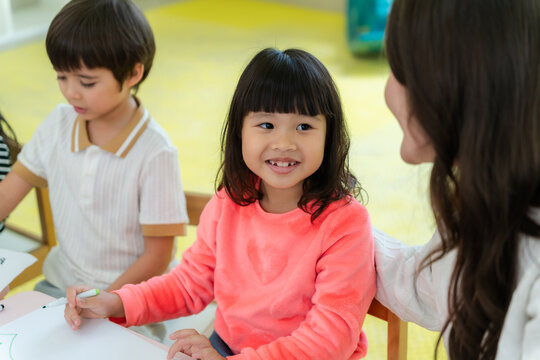 Kind Asian teacher touching shoulder and encouraging a happy little girl during art class at kindergarten.