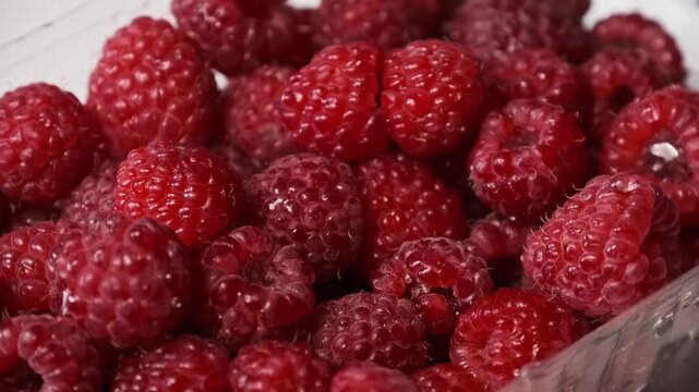 A hand reaches into a clear box to pick a ripe red raspberry from a pile of fresh berries. Concept of healthy eating habits, organic vegetarian nutrition, and sweet summer harvest.