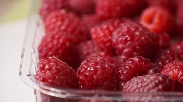 A macro close-up reveals the texture of fresh, ripe red raspberries packed inside a clear plastic container. Concept of healthy snacking, organic farming, and summer harvest freshness.