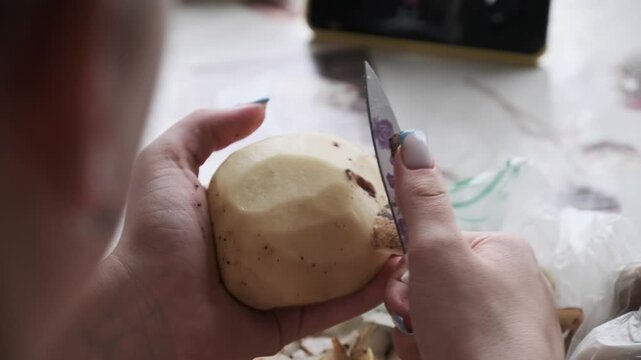 Female hands use a small knife to peel the skin off a raw potato over a kitchen table. Concept of daily household chores, meal preparation, and authentic cooking routines.