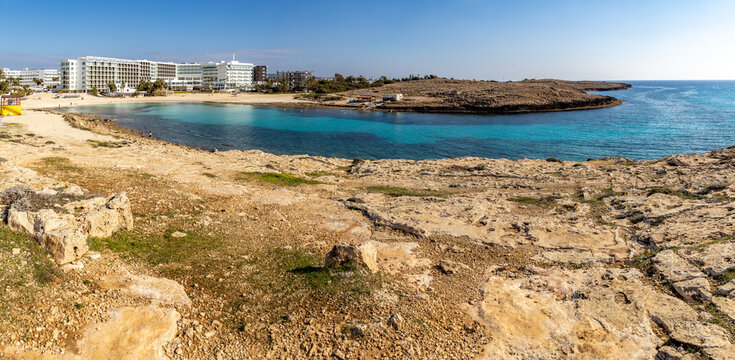 Turquoise water of Vathia Gonia beach bay protected by rocky cape in winter morning. Empty sand coast with white resort hotels during sunny low season, panoramic view, Ayia Napa, Cyprus.