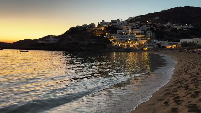 Beautiful beach at sunset in Ios island, Greece. Waves on the sea coast. White architecture on the cliffs. Night lights and their reflections in the water. 
