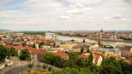 Naklejka premium Aerial panoramic view of Budapest, Hungary showcasing the Danube River, Parliament Building, historic architecture with red-tiled roofs, and city skyline under partly cloudy sky.