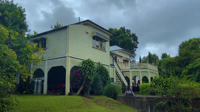 Traditional Queenslander house on stilts surrounded by lush greenery under a cloudy sky.
