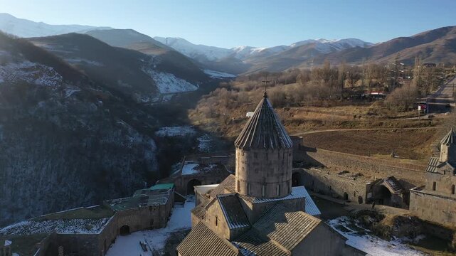 Breathtaking aerial shot of the Tatev Monastery, a medieval Armenian architectural masterpiece perched on a dramatic cliff edge, surrounded by majestic snow covered mountains on a winter day.

