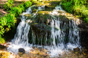Refreshing crystal clear water cascades down mossy rocks in a vibrant forest stream, creating a lively little waterfall surrounded by lush green foliage and sunlit wild nature © PhotoChur