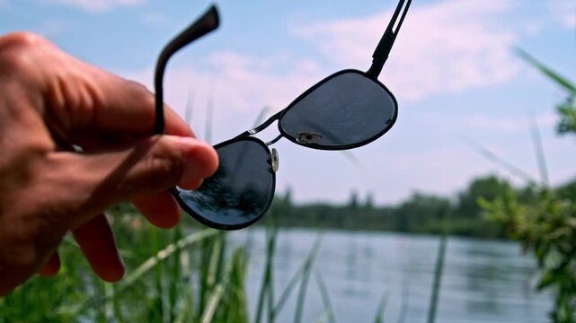 A first person perspective shows a hand holding and moving black aviator sunglasses. The background features a calm lake and bright blue sky creating a relaxed vibe.