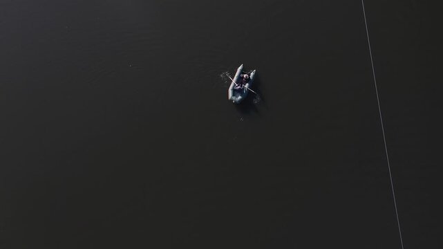 An overhead aerial drone view of a small inflatable boat with two people rowing across calm, dark lake water near a long, straight white safety buoy dividing line.

