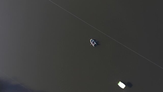 An overhead aerial drone view of a small inflatable boat with two people rowing across calm, dark lake water near a long, straight white safety buoy dividing line.

