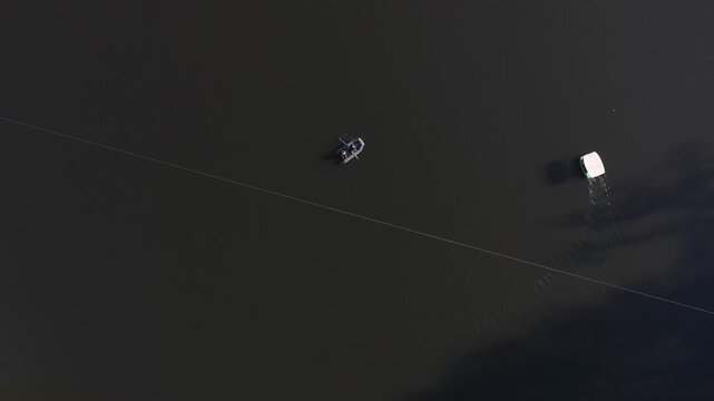 An overhead aerial drone view of a small inflatable boat with two people rowing across calm, dark lake water near a long, straight white safety buoy dividing line.

