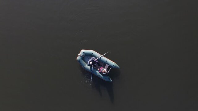 An overhead aerial drone view of a small inflatable boat with two people rowing across calm, dark lake water near a long, straight white safety buoy dividing line.


