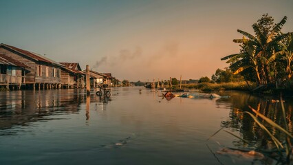 Fototapeta premium Serene Village Scene with Wooden Houses and Palm Trees on Riverside at Sunset